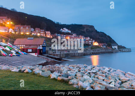 Le luci sono accese nelle case in vista serale del grazioso e pittoresco borgo marinaro, con scogliere, barche & mare calmo - Runswick Bay, North Yorkshire, Inghilterra, Regno Unito. Foto Stock