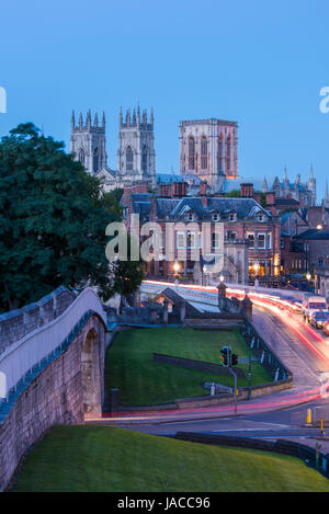 Le mura della città, strada sopra il ponte Lendal & 3 torri iconica del Minster set contro il buio del cielo della sera, incandescenza delle lampade stradali e sentieri di luce - York, GB, UK. Foto Stock