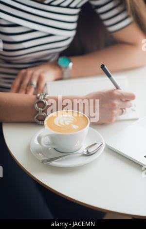Ritratto di close-up di mani rendendo note su carta, tazza di cappucciono e laptop in piedi su un tavolo in un bar. Foto Stock