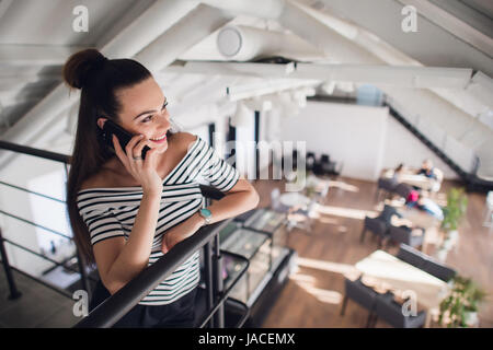Bella donna adulta in camicia classica sta parlando al telefono cellulare, guardando lontano e sorridenti mentre sta in piedi vicino al suo posto di lavoro. Foto Stock