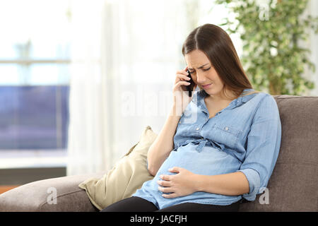 Giovane donna incinta soffre il mal di pancia e chiamando al telefono seduto su un divano nel soggiorno in un interno di una casa Foto Stock