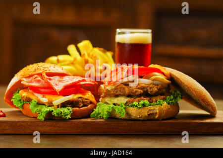 Close up di due deliziosi hamburger con carne di manzo, pancetta, cipolla, pomodoro, lattuga e formaggio con una offuscata un bicchiere di birra e patatine fritte dietro sulla tavola di legno Foto Stock