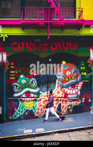 Una donna che cammina verso il basso un vicolo verso il basso oltre una parete di graffiti raffiguranti dragone cinese dancing in Chinatown di San Francisco, California Foto Stock