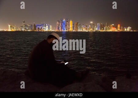 Un uomo si siede sulla parete del mare nel porto Dhow guardando al suo telefono mobile. In fondo è la vista su tutta la baia verso l'alto di business Foto Stock