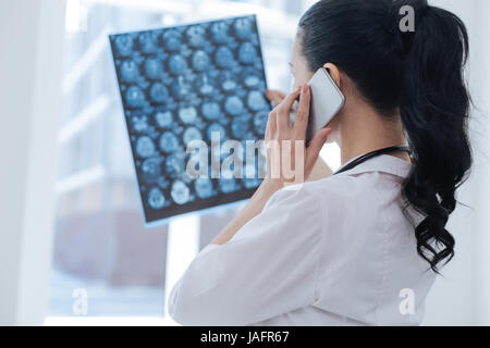 Il controllo della salute delle modifiche. Le figure professionali coinvolte oncologo femmina lavorando al roentgen armadio mentre esaminando scansione cerebrale e di immagine utilizzando smartpho Foto Stock