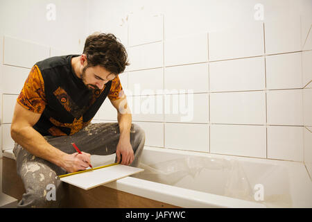 Un builder, installatore lavora in un bagno, segnando una piastrella con una matita. Foto Stock