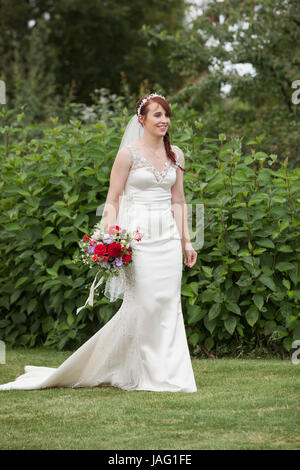 Sposa nel suo abito da sposa in piedi in un giardino, tenendo wedding bouquet di fiori di colore rosso. Foto Stock