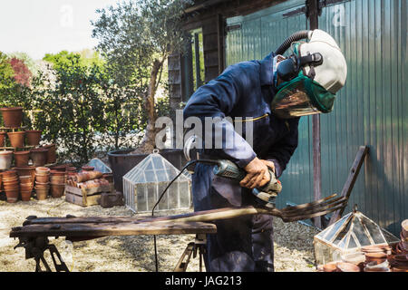 Uomo in piedi all'aperto, indossare una maschera per il viso, lavorando su un metallo forcone con una smerigliatrice angolare. Foto Stock