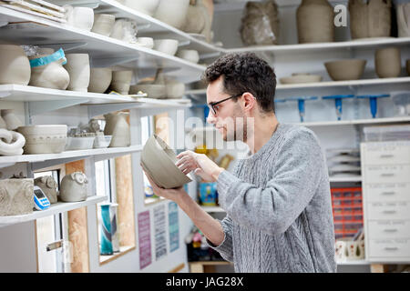 Un uomo di ispezione di una pentola di creta, prima della cottura. Mensole in ceramica studio pieno di pentole, lavori in corso. Foto Stock