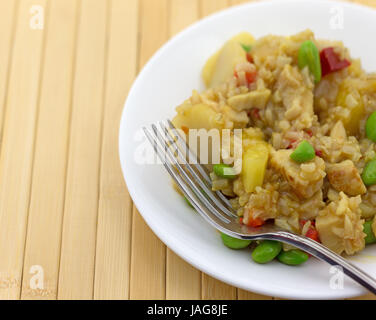 Un pasto di pollo, riso, acqua di castagne, peperoni rossi e pezzi di ananas in una salsa con una forcella su una piastra bianca sulla cima di un legno posto mat. Foto Stock