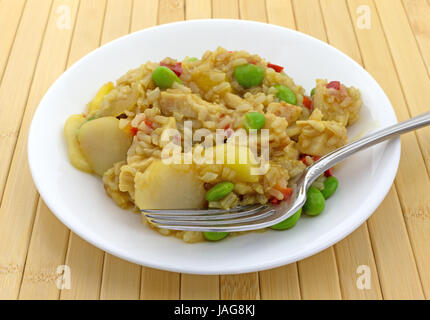 Un pasto di pollo, riso, acqua di castagne, peperoni rossi e pezzi di ananas in una salsa con una forcella su una piastra bianca sulla cima di un legno posto mat. Foto Stock