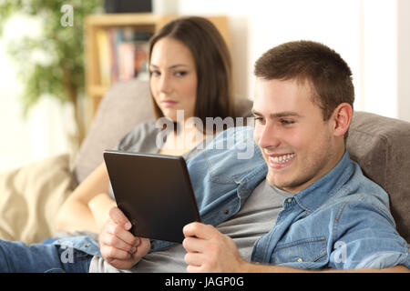 Il marito guardando il contenuto online di un tablet e ignorando la sua moglie arrabbiato che è seduto accanto a lui su un divano a casa Foto Stock