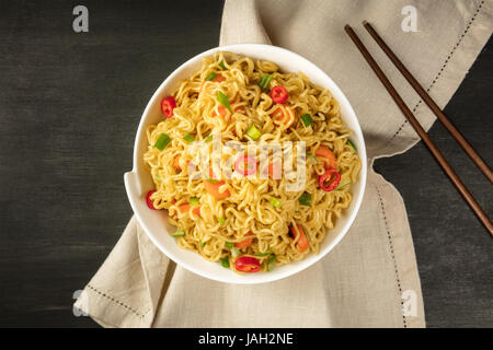 Una ciotola di instant noodle cinesi con le cipolle verdi, Red Hot Chili Peppers, e carote, ripresa dall'alto su una texture scuro con bacchette e una plac Foto Stock