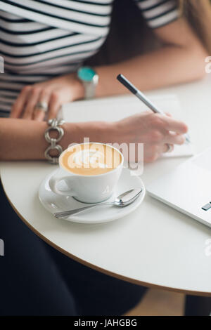Close-up le mani della donna in possesso di una penna sul notebook vuota con la tazza di caffè accanto ad essa. Vista superiore della composizione. Luogo di lavoro della femmina di office Foto Stock