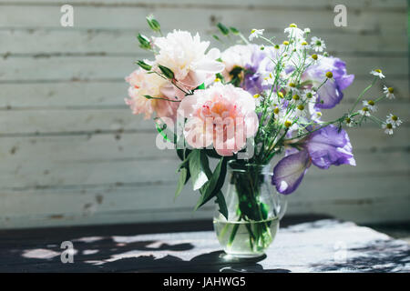 Bouquet di peonie bianco, chamomiles e iride fiori in vaso di vetro. Estate sfondo. Foto oscurata Foto Stock