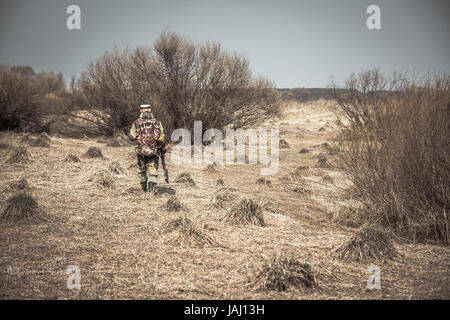 Hunter uomo in camuffamento con la pistola passando attraverso la zona rurale con erba secca e cespugli durante la caccia Foto Stock