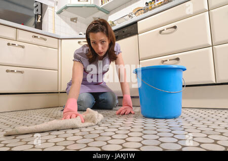 Erwachsene Hausfrau kniet in ihrer hellen Einbauküche und reinigt sauer frustriert und den Fußboden mit Eimer und Putzlappen. Foto Stock