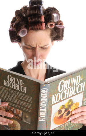 Erwachsene Hausfrau mit Lockenwicklern in den Haaren und einem Bademantel liest in einem großen Kochbuch vor weißem Hintergrund. Foto Stock