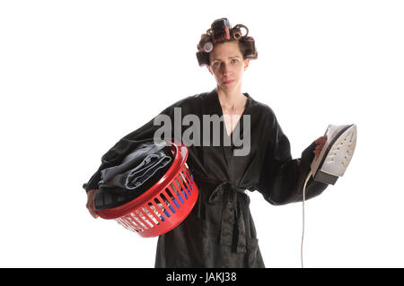 Erwachsene Hausfrau mit Lockenwicklern in den Haaren und einem Bademantel steht vor weißem Hintergrund und Mit einem Korb Bügelwäsche und einem Bügeleisen. Foto Stock