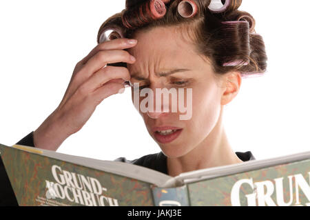 Erwachsene Hausfrau mit Lockenwicklern in den Haaren und einem Bademantel liest in einem großen Kochbuch vor weißem Hintergrund. Foto Stock