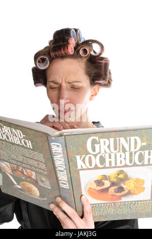 Erwachsene Hausfrau mit Lockenwicklern in den Haaren und einem Bademantel liest in einem großen Kochbuch vor weißem Hintergrund. Foto Stock