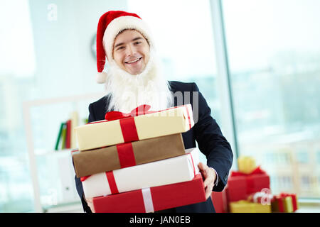 Immagine di happy businessman in Santa Cappello e barba holding pila di doni e guardando la telecamera in ufficio Foto Stock