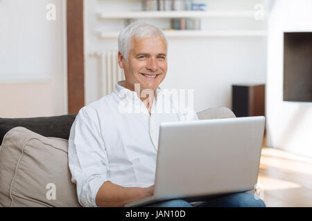 Sorridente uomo maturo seduto su un divano digitando su un computer portatile in una stanza vivente Foto Stock