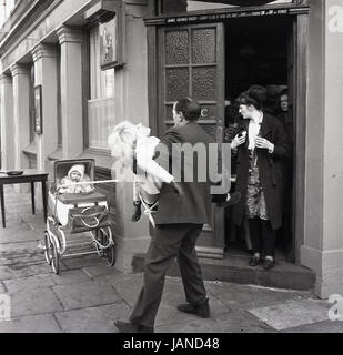 1965, storico, immagine mostra un uomo che porta una signora al di fuori dell'entrata di una casa pubblica che è collassato in esaurimento dopo la concorrenza nell'annuale gara di pancake giù Old Kent Road, Peckham, Londra. Si noti che un bambino nella PRAM è stato lasciato sulla propria al di fuori del pub. Foto Stock