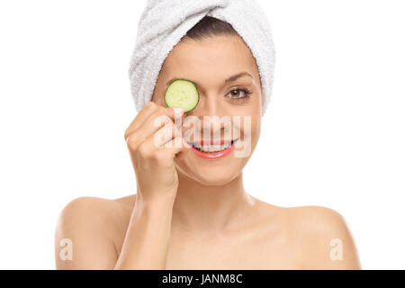 Giovane donna tenendo una fetta di cetriolo sopra il suo occhio e sorridente isolati su sfondo bianco Foto Stock