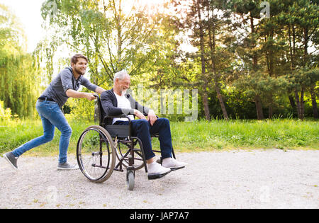 Hipster figlio in esecuzione con padre disabili in sedia a rotelle al parco. Foto Stock