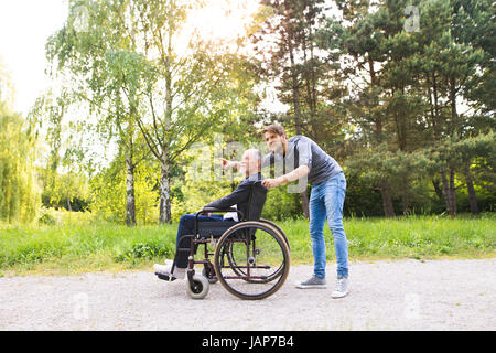 Hipster figlio a piedi con padre disabili in sedia a rotelle al parco. Foto Stock