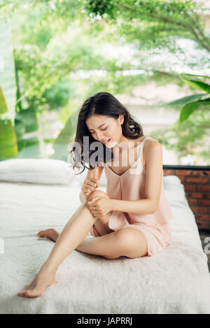 Giovane e bella donna che soffrono di dolore al ginocchio mentre è seduto sul letto di casa Foto Stock