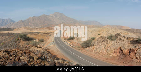 Vista panoramica di una strada che si snoda attraverso l'arido paesaggio wadi dell'Oman con aspre montagne sotto un cielo azzurro e limpido, viaggio avventuroso in Medio Oriente Foto Stock