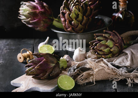 Non cotti interi umido organico viola i carciofi in ciotola vintage e su legno tagliere con calce, aglio, filo, tessuti cilicio su legno scuro Foto Stock