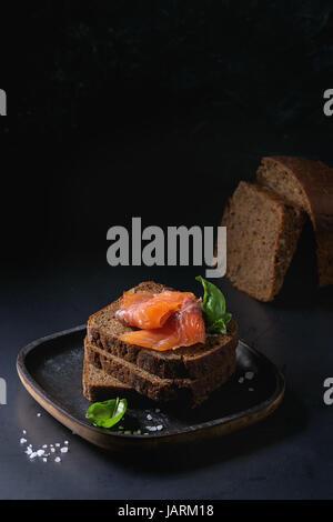 Pila di casalinga a fette di pane di segale con salmone affumicato, sale marino e basilico fresco sul piatto di legno scuro su sfondo nero. Foto Stock