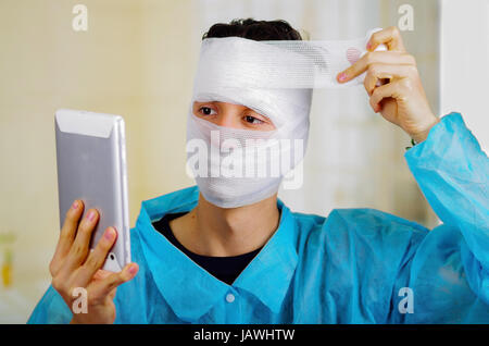 Ritratto di un giovane uomo con trauma nella sua testa e bendato elastico intorno alla sua testa portante un tablet. Foto Stock