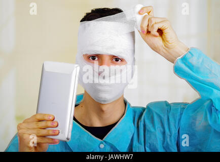 Ritratto di un giovane uomo con trauma nella sua testa e bendato elastico intorno alla sua testa portante un tablet. Foto Stock