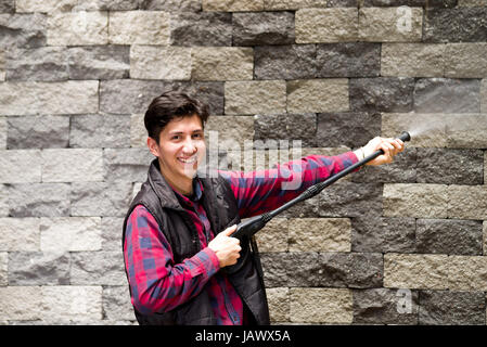 Bel giovane uomo che indossa disegno quadrato rosso tenendo alta pressione pistola ad acqua, puntando verso il grigio muro di mattoni Foto Stock