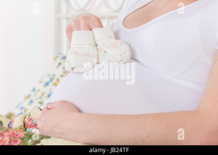 Donna incinta con bambini's bootees siede su un letto in camera. Foto Stock