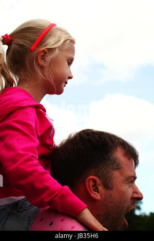 Piccola ragazza seduta sul suo padre la spalla con le nuvole in background e papà sbadigliare Foto Stock