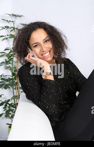 Junge, hübsche Frau sitzt auf dem Sofa und telefoniert Mit einem Smartphone Foto Stock