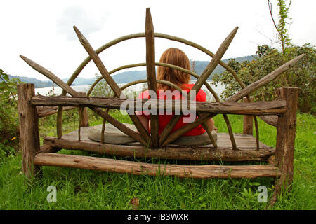 Una donna si ferma a poggiare su un grande banco di legno nel bosco su una riva del lago Foto Stock