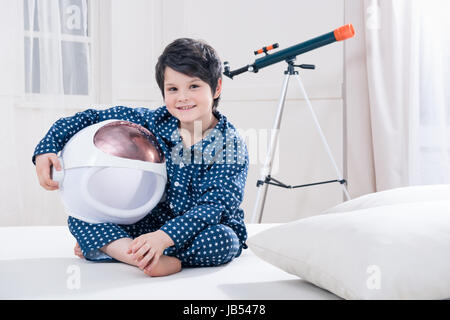 Carino piccolo ragazzo in pigiama spazio di contenimento casco seduta sul letto Foto Stock