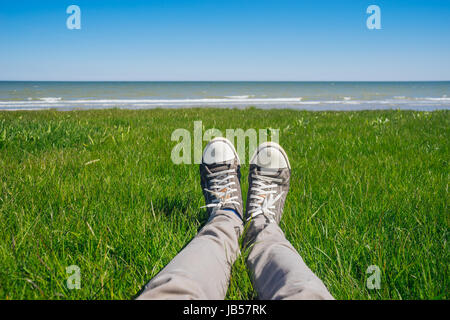 Gambe maschio in jeans e scarpe da tennis su erba fresca contro seacoast. Giorno d'estate e di sole Foto Stock