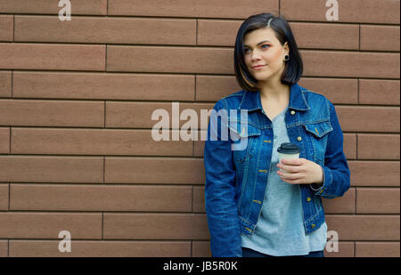 Bella bruna giovane donna con gallerie di orecchie in un jeans blu giacca con una tazza di caffè in piedi di fronte a un muro di mattoni. Foto Stock