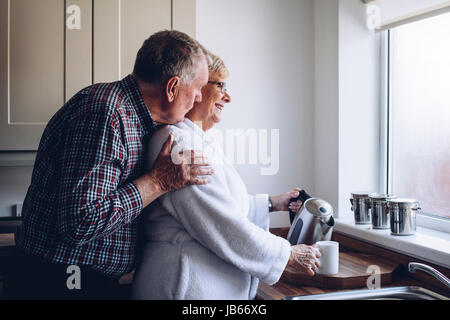 Senior uomo baciare sua moglie da dietro mentre lei versa una tazza di tè. Foto Stock