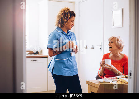 Home operatore sanitario a parlare al suo paziente nella sua cucina come loro bere il tè e mangiare un pan di spagna. Foto Stock