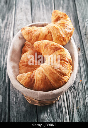 Angolo di alta vista di pane appena sfornato a scaglie golden croissant in un cestello servita su un vecchio rustico tavolo in legno con una texture spiovente Foto Stock