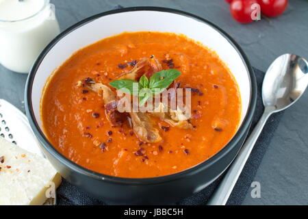 Spesso la zuppa di pomodoro con basilico e pancetta fritta in un nero vaso in ceramica su un grigio Sfondo astratto. Mangiare sano concetto Foto Stock