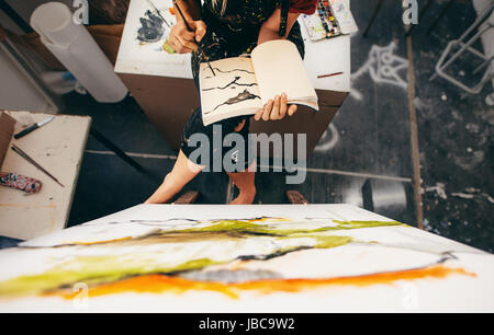 Vista superiore del pittore femmina seduto in studio la realizzazione di un disegno nel suo libro. Ritagliato shot donna artista pittura nel suo libro. Foto Stock